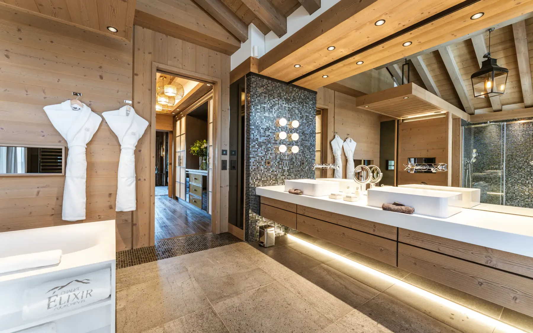 Luxurious bathroom with wooden decor, double sinks, large mirror, hanging robes, and soft lighting, creating a warm and inviting atmosphere.