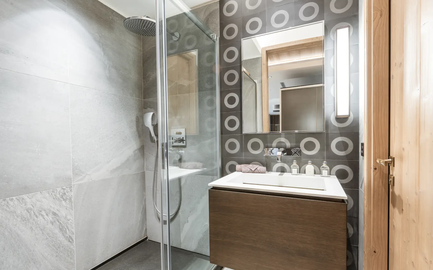 Modern bathroom with glass shower, circular patterned tiles, wooden vanity, rectangular mirror, and soft lighting.