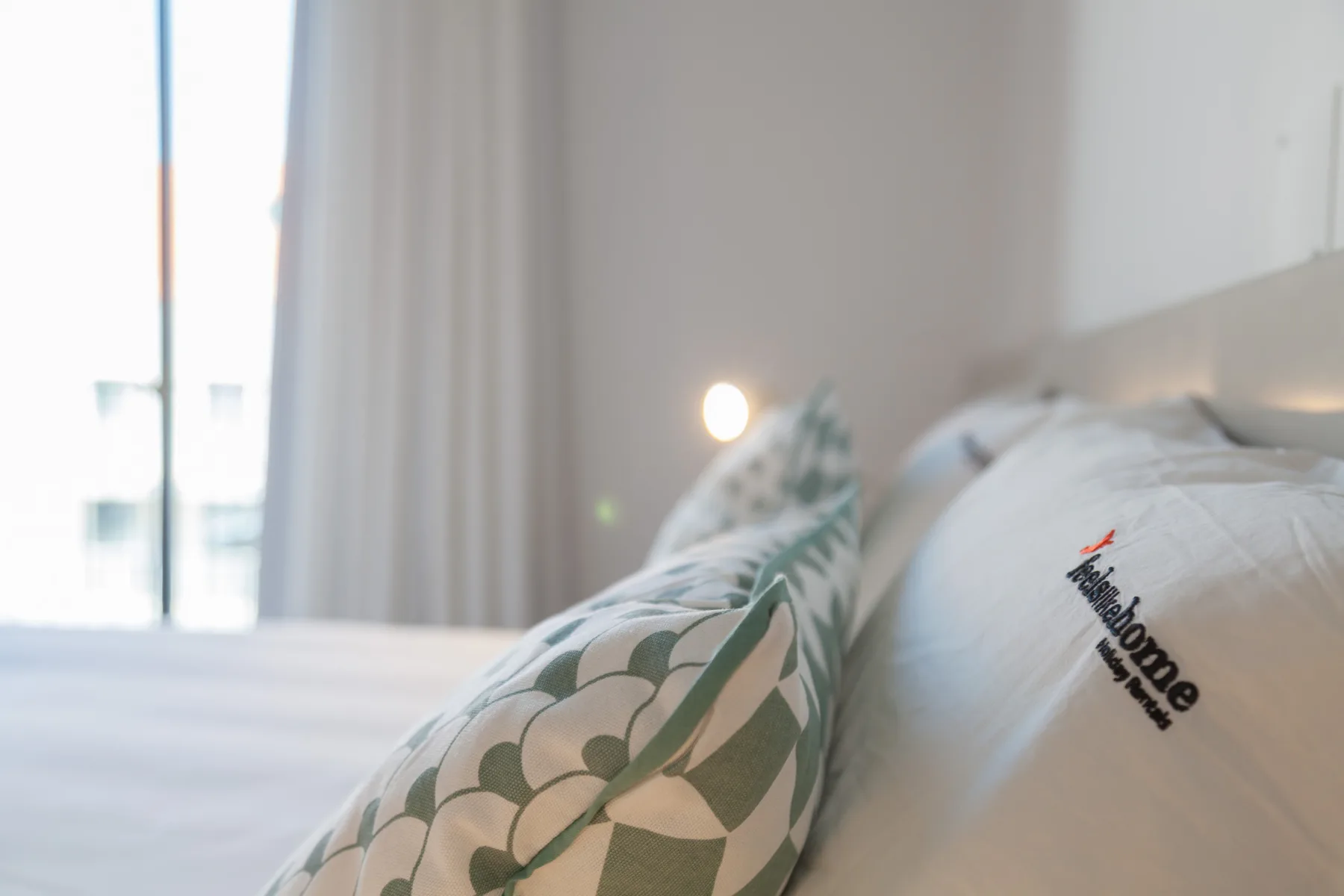 A bedroom with a patterned cushion on the bed beside a bright window.
