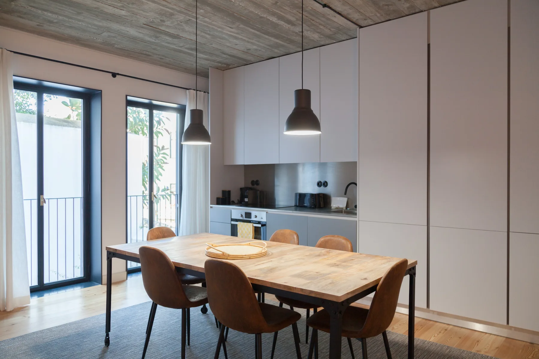 A dining area with a wooden table and kitchen cabinets beside tall windows.