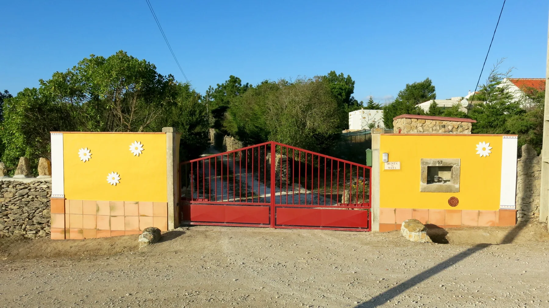 A red metal gate stands between yellow entrance walls beside a gravel road.