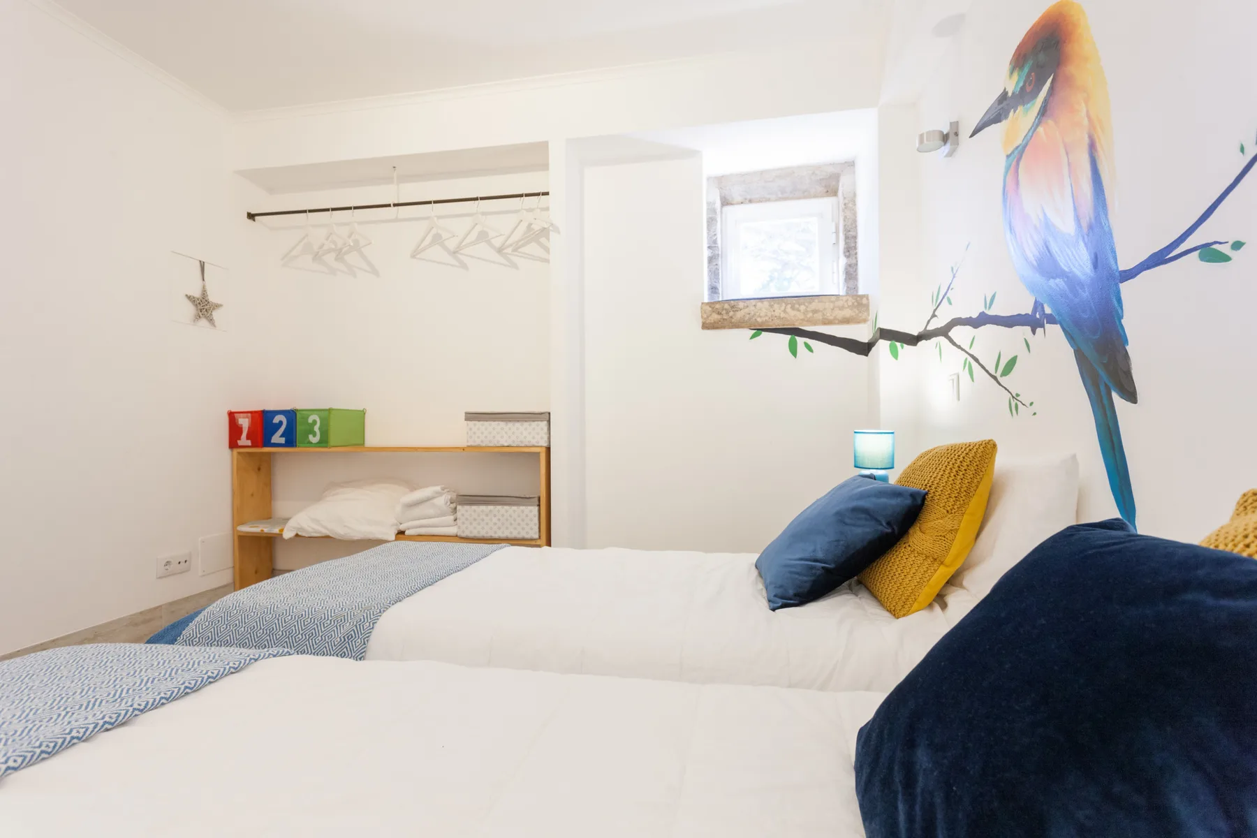A bright bedroom with white bedding, blue pillows, and a colorful bird mural.