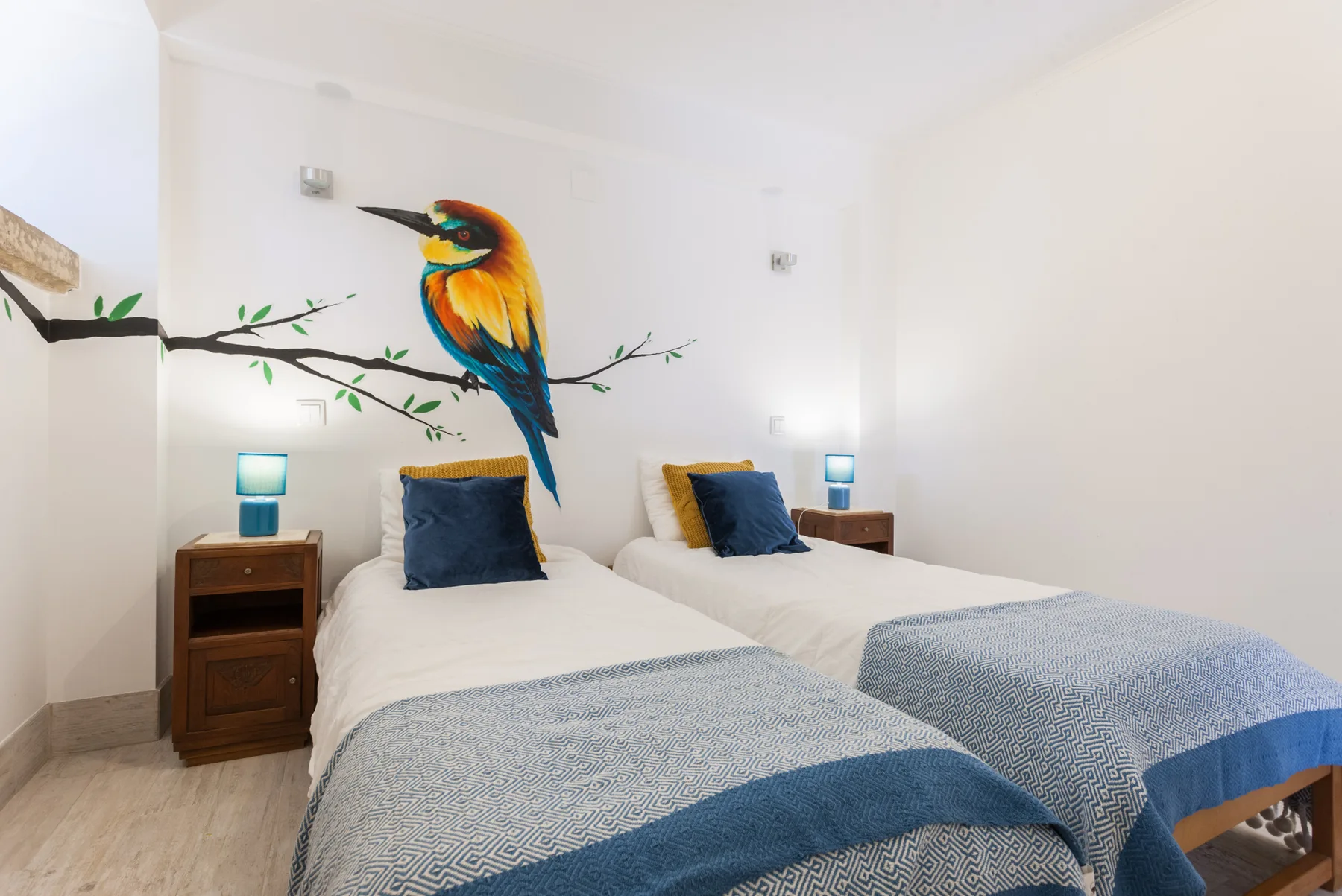 A bedroom with two beds and a colorful bird mural on the wall.