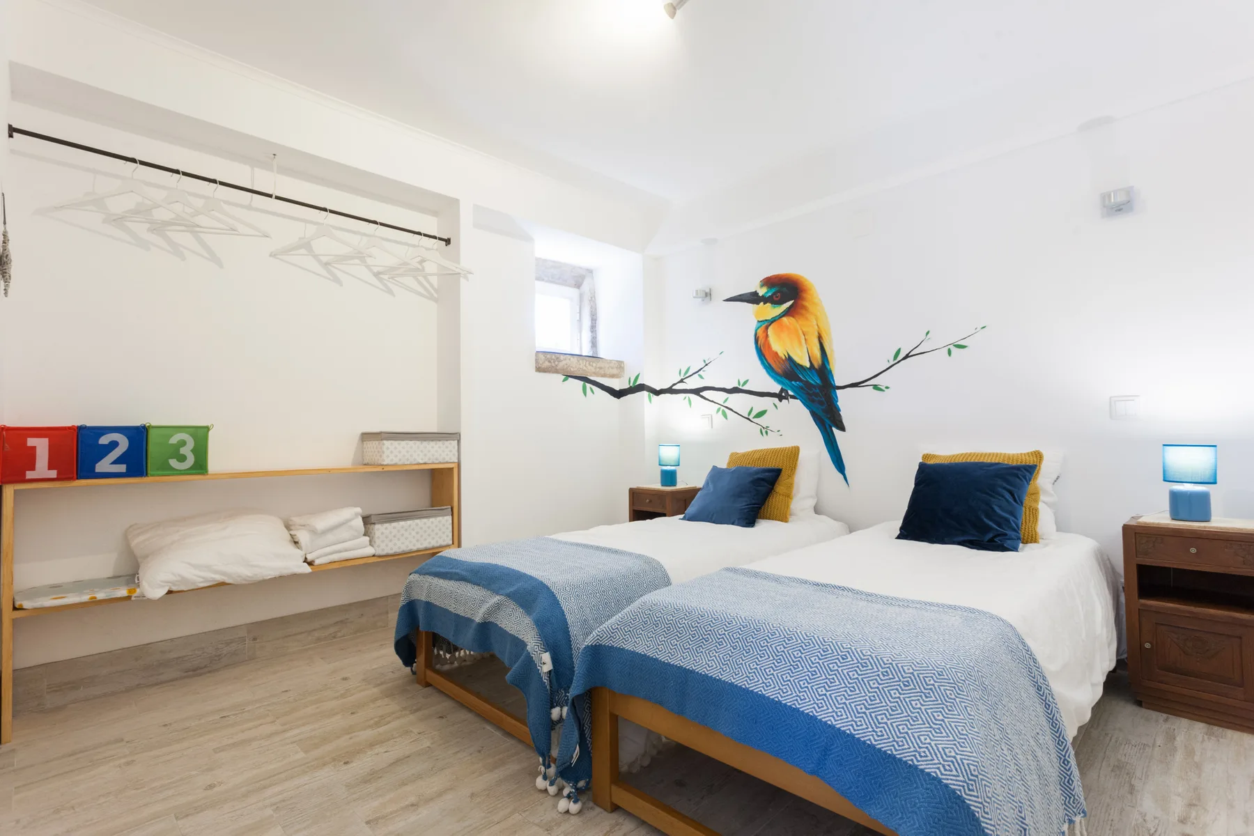 A bright bedroom with two beds, blue bedding, and a colorful bird mural.
