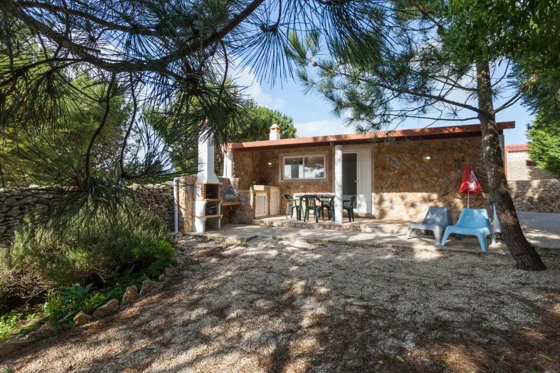 A stone house with outdoor seating sits beneath pine trees.