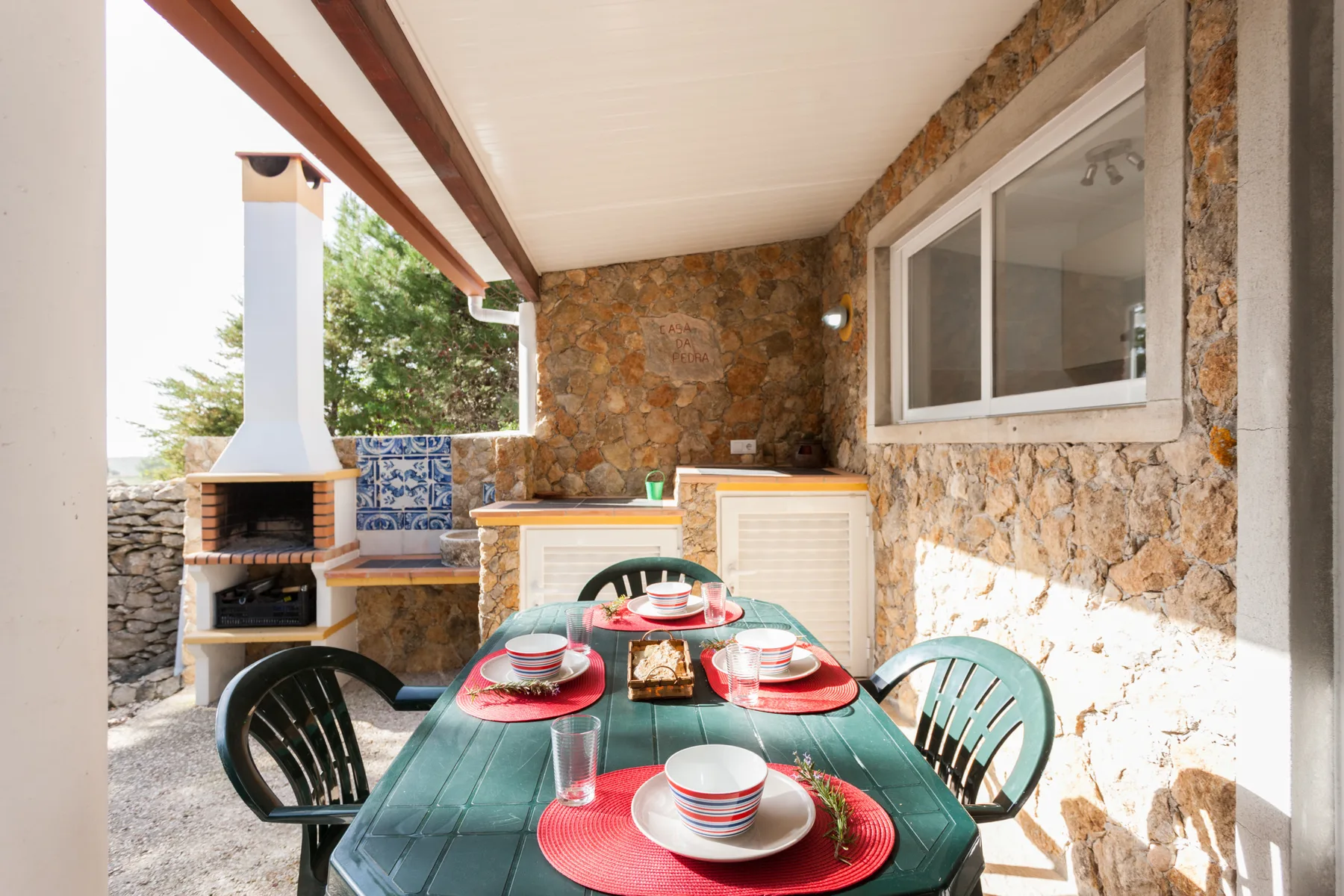 Covered outdoor dining area with a table set beside a stone wall and barbecue.