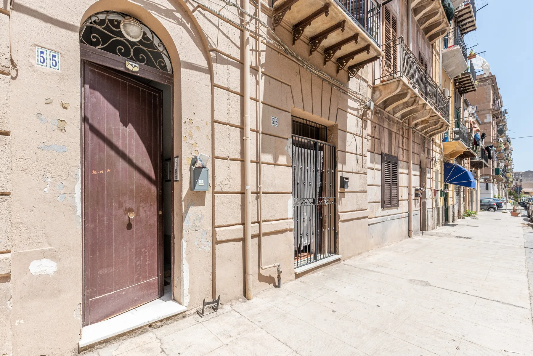 Una calle soleada bordeada de edificios claros, balcones y una puerta oscura de entrada.