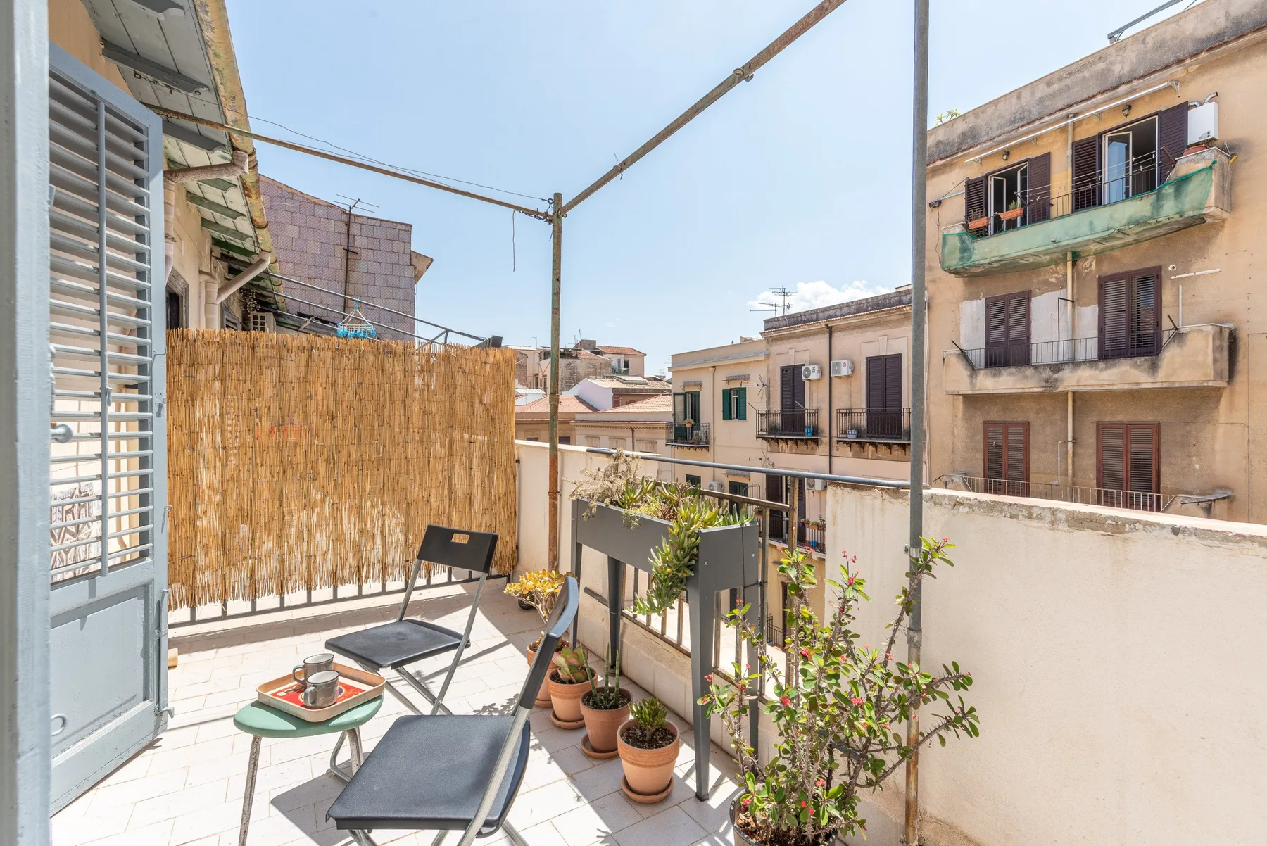 Una terraza soleada con asientos al aire libre, plantas en macetas y edificios vecinos a la vista.