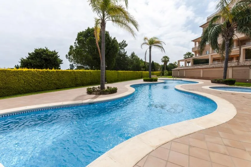 Una piscina exterior curva junto a una terraza pavimentada y palmeras.