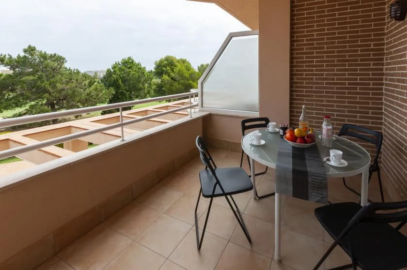 A balcony dining area with a glass table and seating overlooks trees.