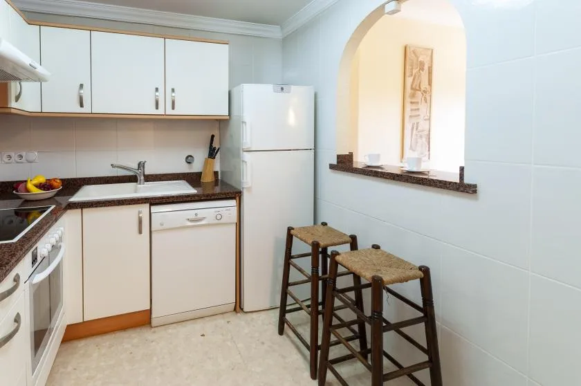 Una cocina compacta con gabinetes blancos, un refrigerador y dos taburetes junto a la barra.