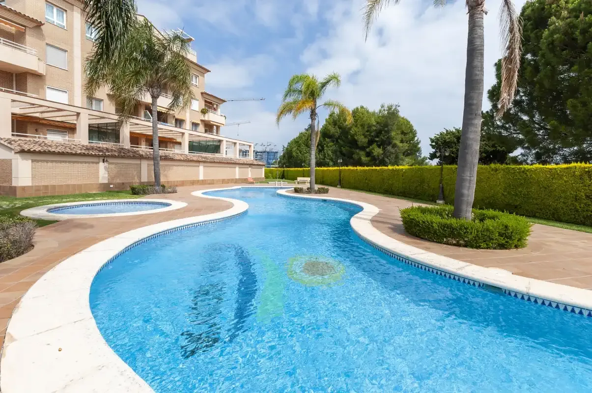 Una piscina exterior curvada está junto a un jardín ajardinado y un edificio de apartamentos.