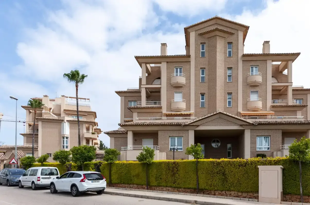 Un edificio residencial de varias plantas se alza tras un seto recortado junto a una calle.
