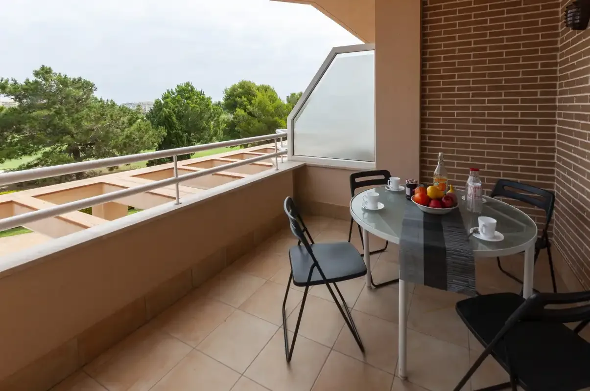 Un balcón embaldosado con una mesa de vidrio y asientos da a los árboles.