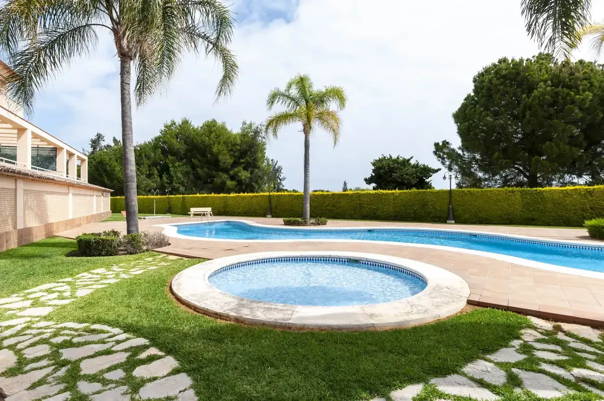 Una piscina exterior y un jacuzzi circular están rodeados de palmeras, senderos y un seto recortado.