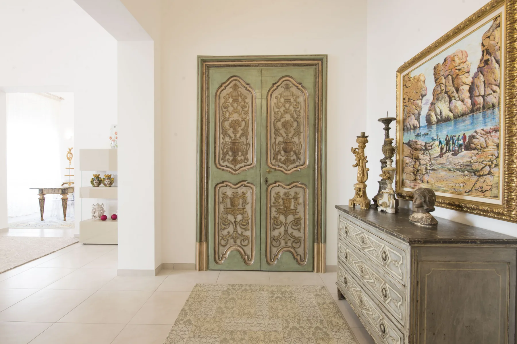 Un pasillo interior tiene una puerta doble ornamentada, un mueble tallado y una obra enmarcada.