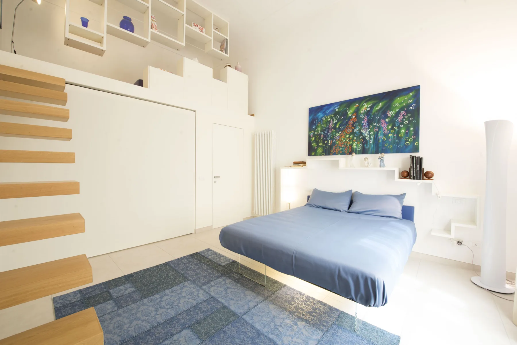 Un dormitorio con una cama azul, arte colorido en la pared y una alfombra estampada.