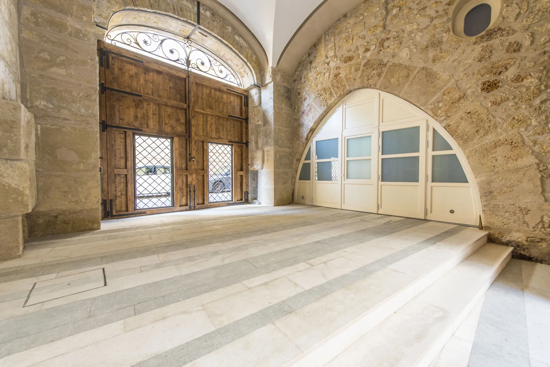 Un vestíbulo interior de piedra con puertas de madera arqueadas y amplios escalones claros.