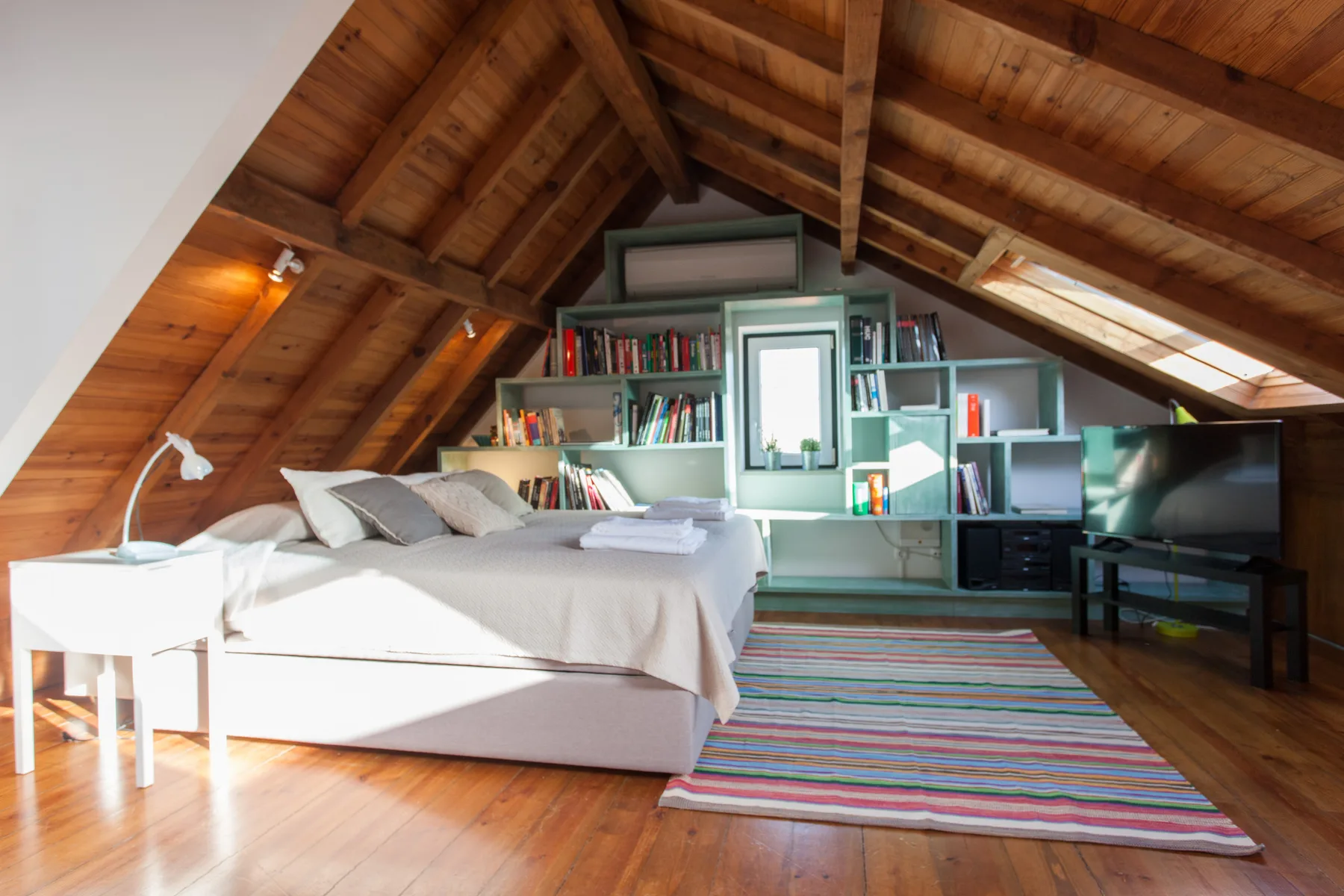 A bedroom with a bed, bookshelves, and a colorful striped rug beneath a wooden ceiling.