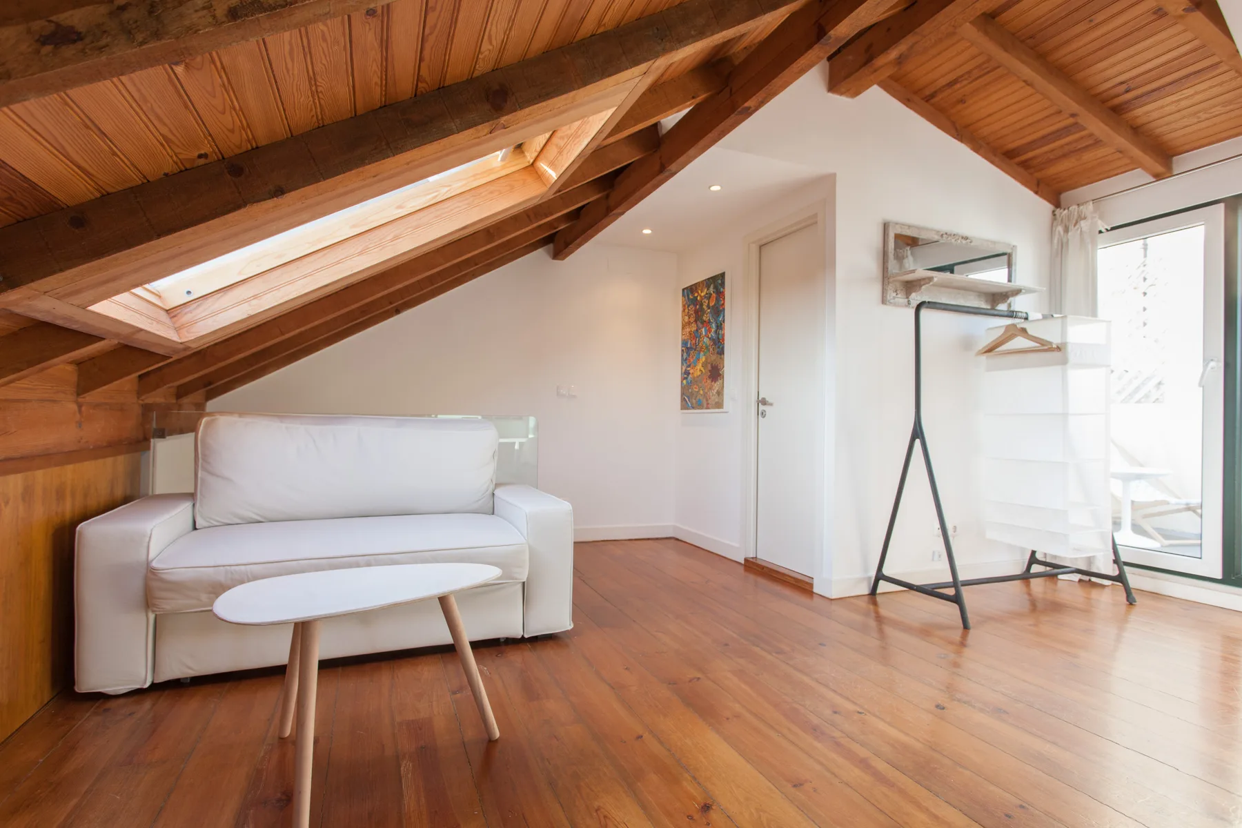 A bright attic living area with seating and a clothing rack beside glass doors.