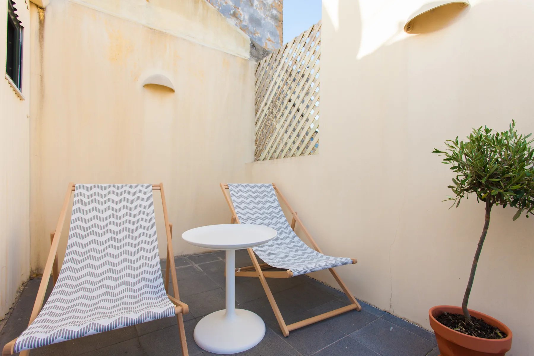 A sunny terrace with two lounge chairs, a small table, and a potted tree.