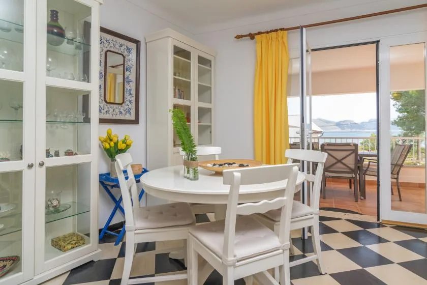 A bright dining area with a round table, white seating, and glass doors opening onto a balcony.