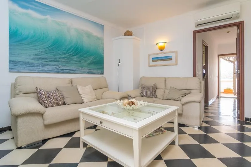 A bright living room with beige seating, a glass coffee table, and a large ocean-wave artwork.