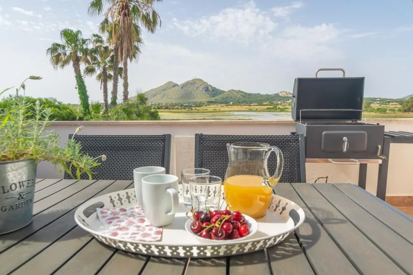 An outdoor patio table holds breakfast drinks and fruit, with a barbecue and palm trees nearby.