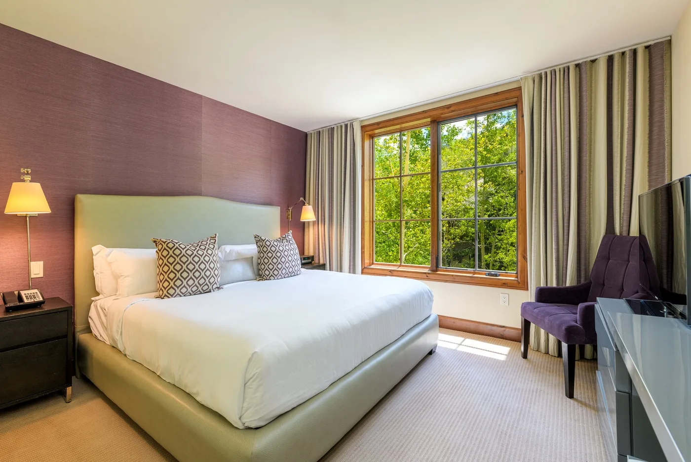 Un dormitorio con una cama bien hecha junto a una gran ventana con vista a árboles frondosos.