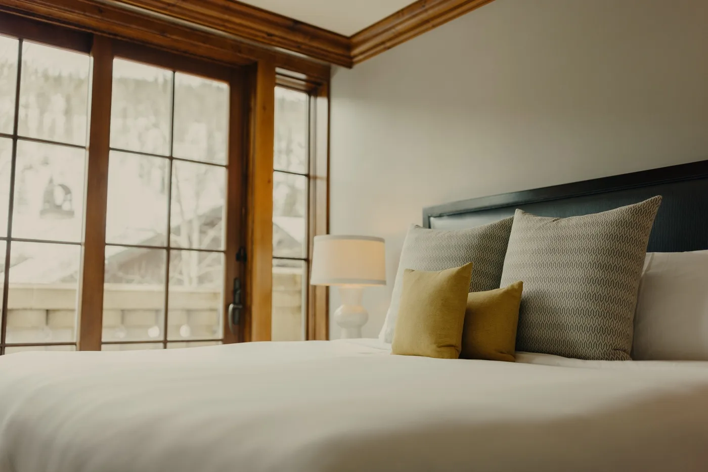 Un dormitorio con una cama, almohadas neutras y ventanas altas con marcos de madera.