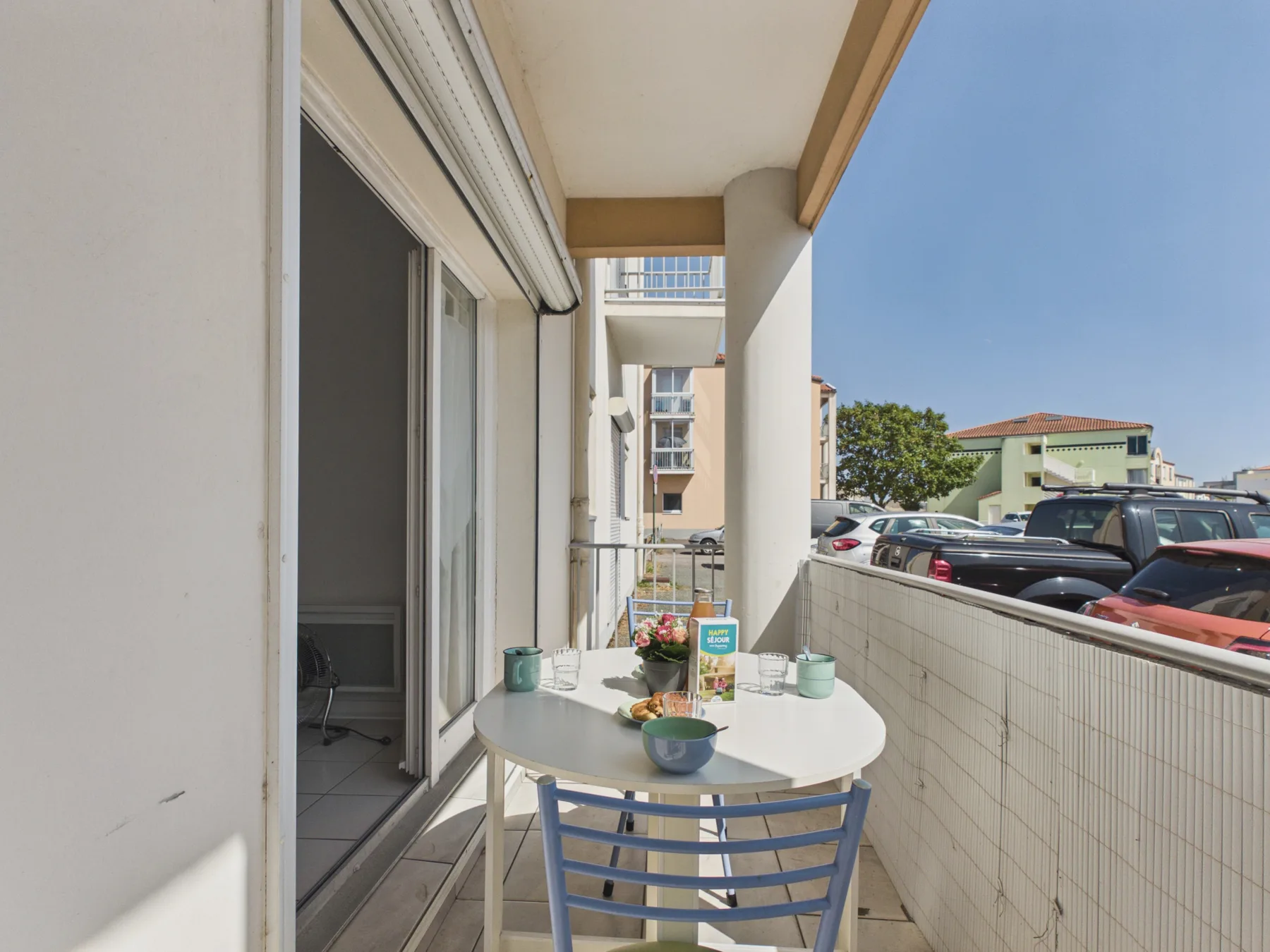 A narrow outdoor balcony with a small table and seating overlooks a parking area.