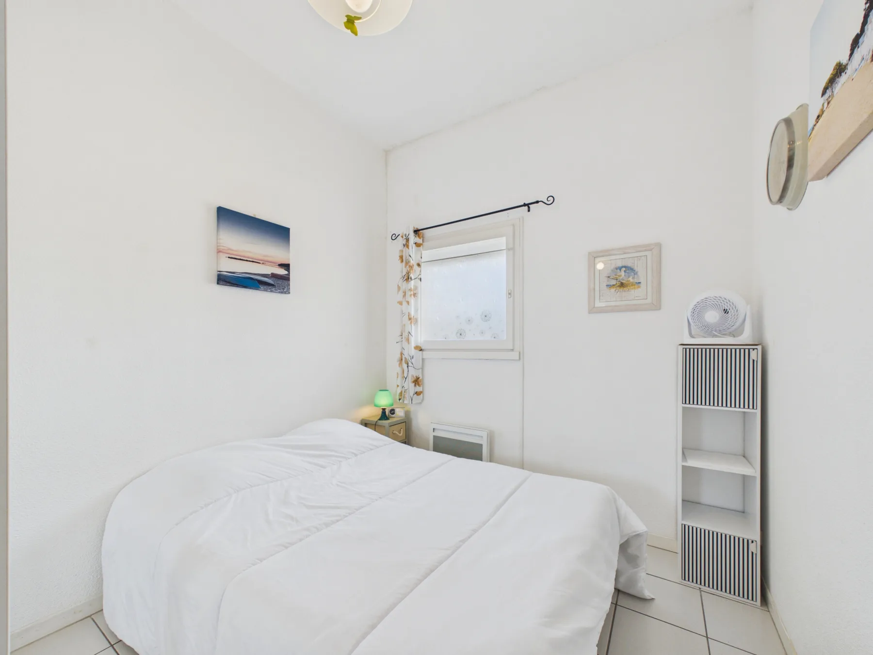 A bright bedroom with a bed, a window, and a narrow shelving unit.