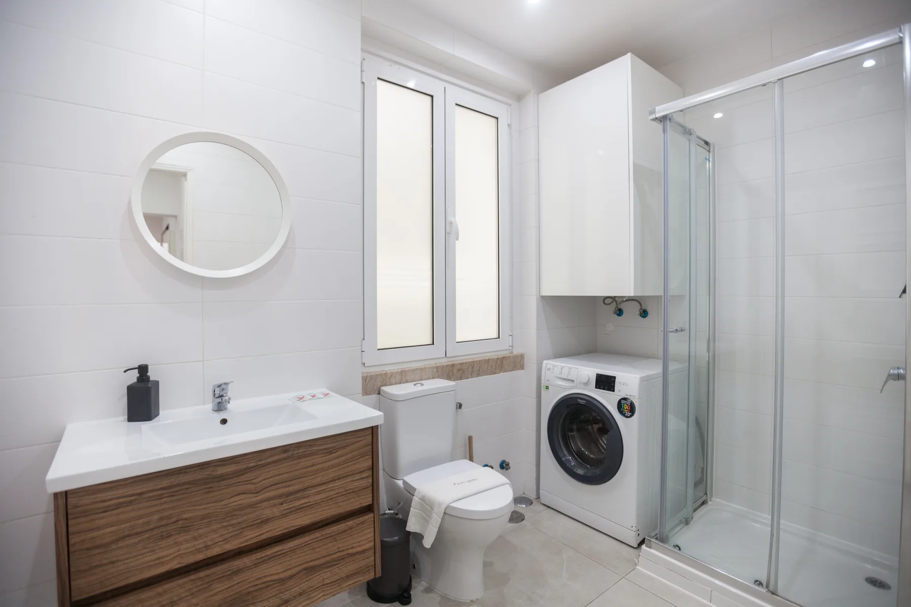 A bright bathroom with a vanity, toilet, washing machine, and shower.