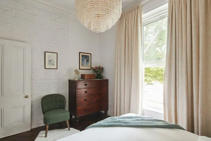 A bedroom with a bed, a wooden dresser, and tall curtained windows.