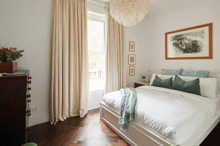 A bedroom with a white bed, pale curtains, and framed artwork.