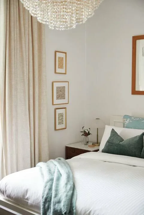 A bright bedroom with a neatly made bed, pale curtains, and framed artwork.