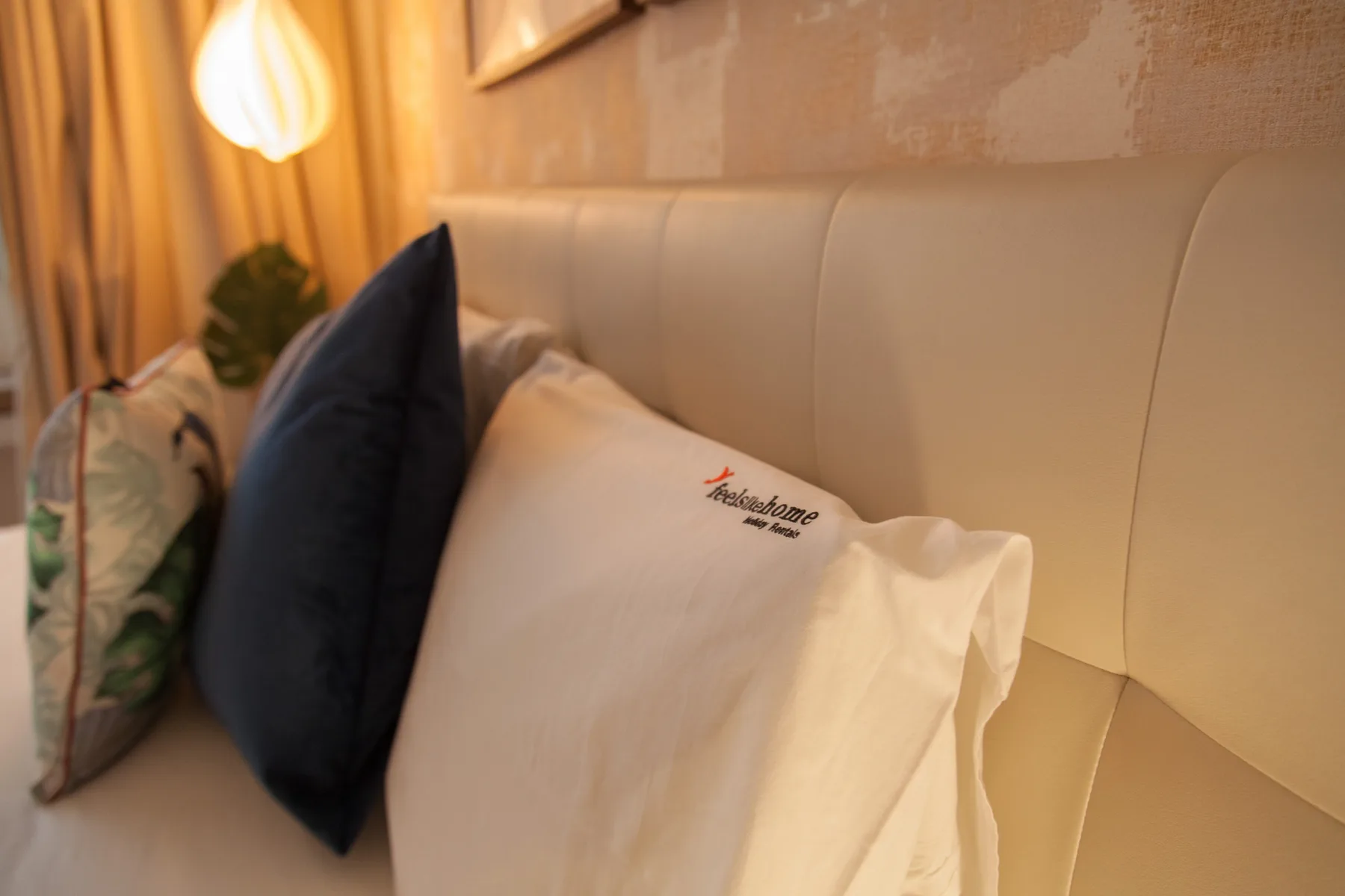 Una cama de dormitorio con varias almohadas junto a una lámpara cálida encendida.