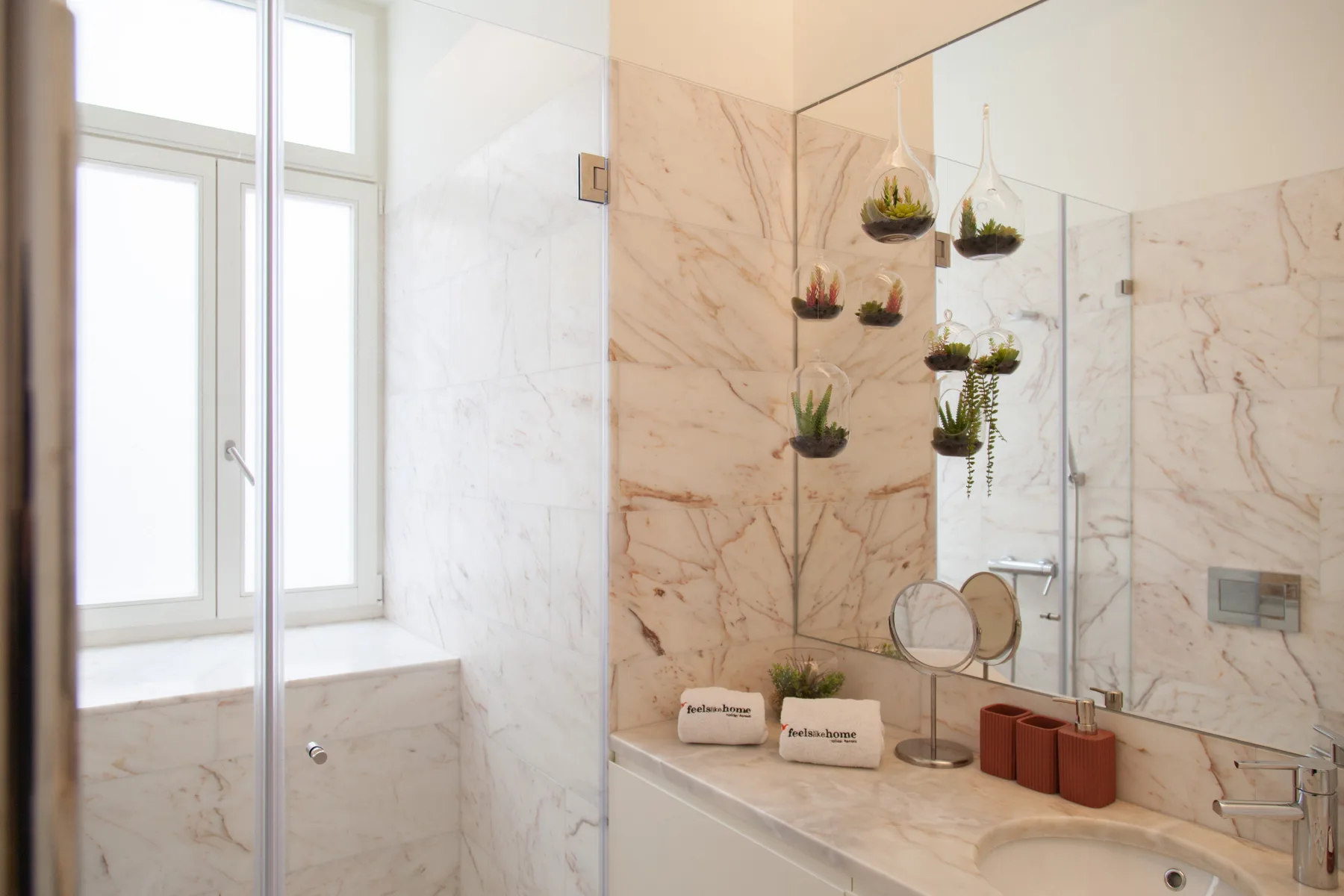 Un baño luminoso de mármol con bañera, mampara de ducha de vidrio y plantas colgantes.