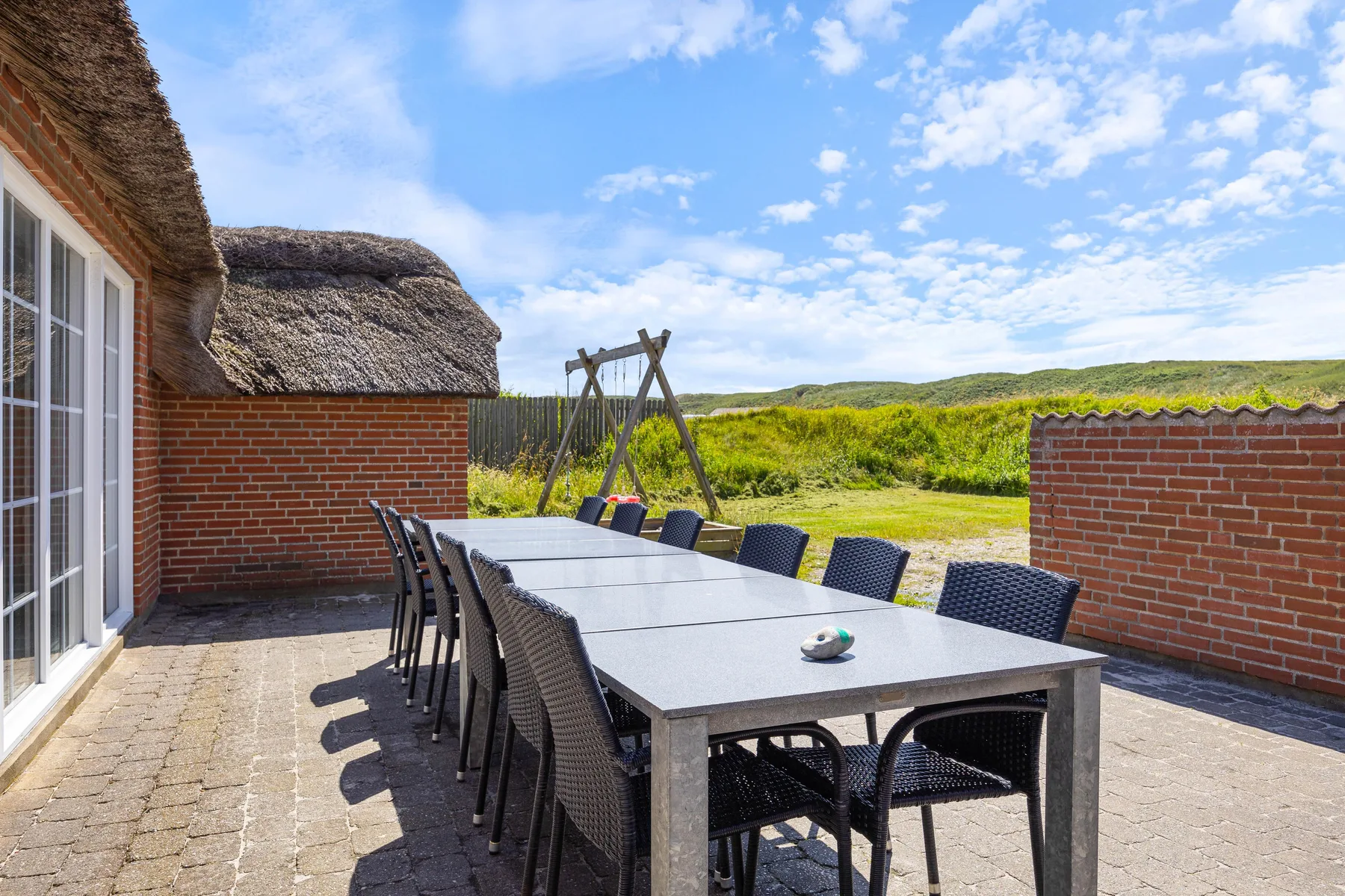 An outdoor dining area with a long table and dark seating beside a brick wall.