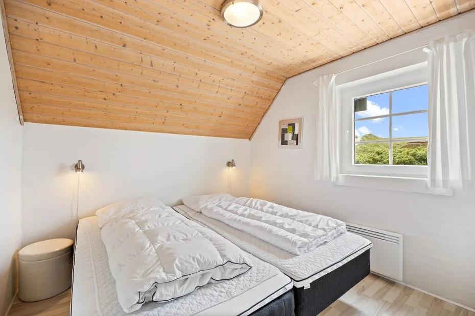 A bright bedroom with two beds beneath a sloped wooden ceiling.