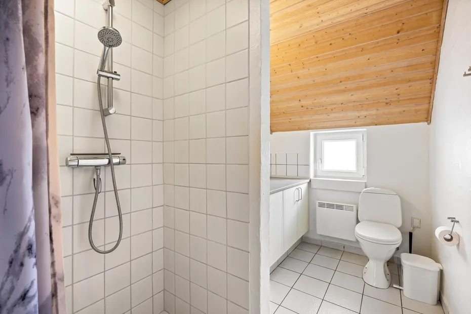 A tiled bathroom with a shower and a toilet beneath a sloped wooden ceiling.