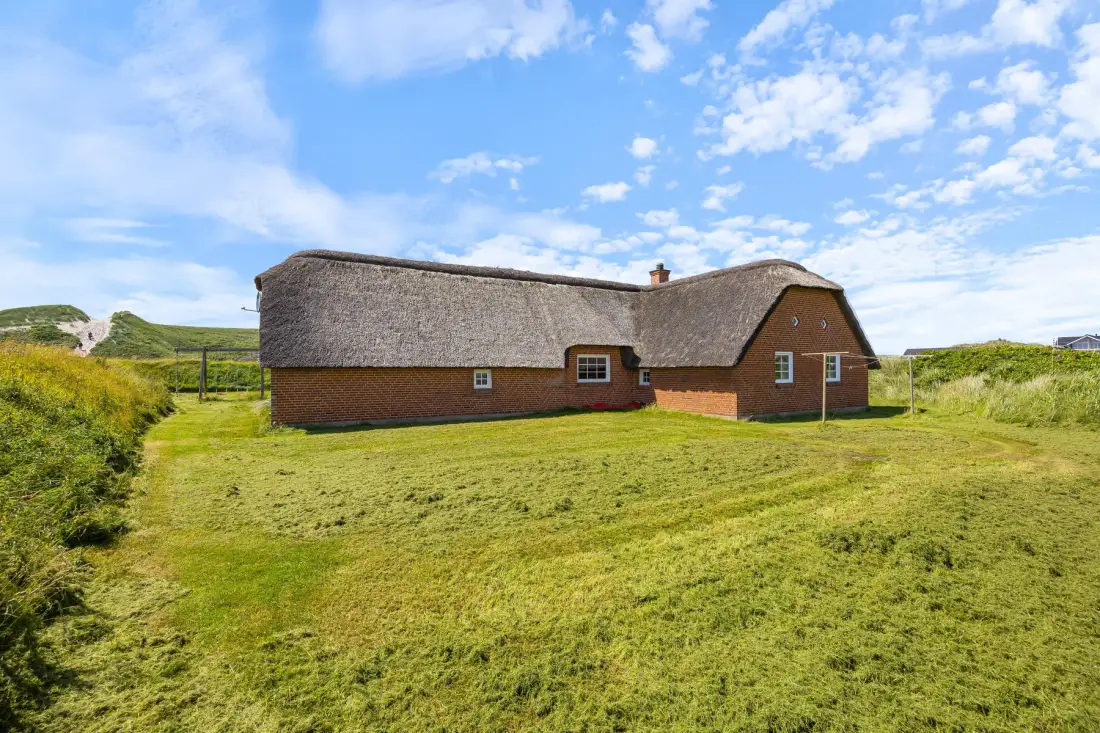 A thatched house stands in a grassy landscape beneath a blue, cloud-dotted sky.