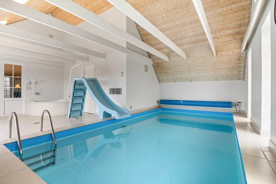 An indoor swimming pool with a blue slide beneath a sloped wooden ceiling.