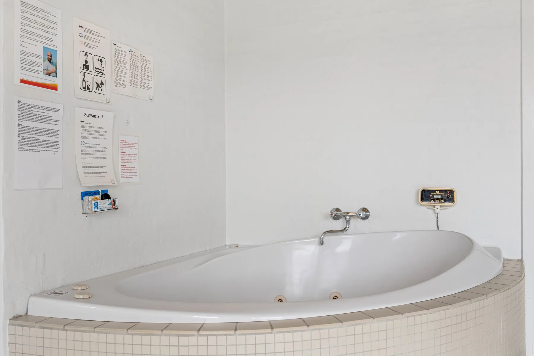 A built-in bathtub sits beneath several notices on a white bathroom wall.