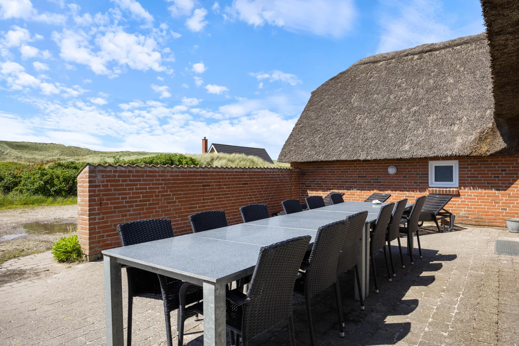 An outdoor dining area with a long table and several chairs beside a brick building.