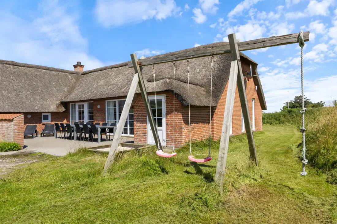 A brick house with a thatched roof stands behind a wooden swing set on the lawn.