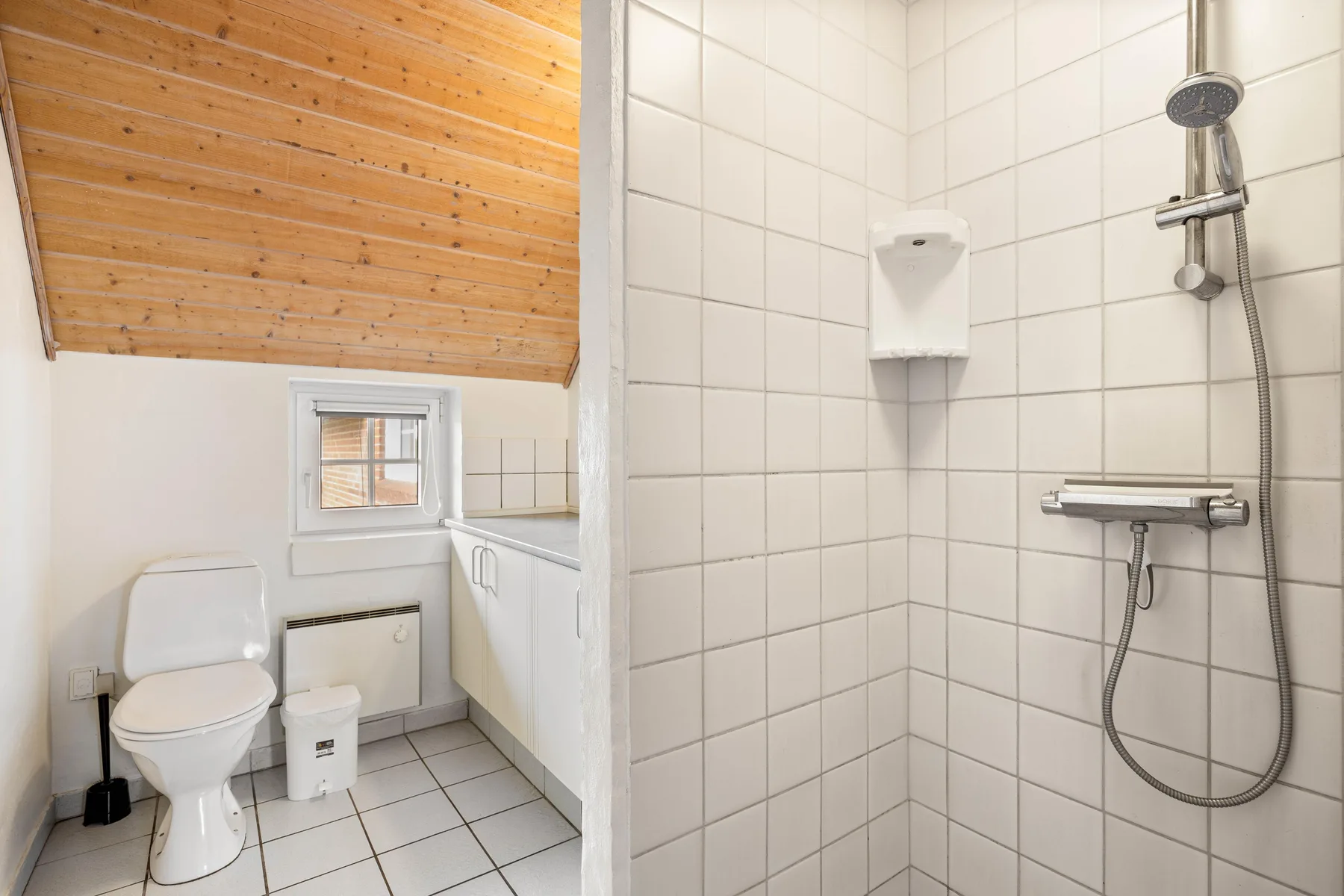 A tiled bathroom with a shower, toilet, and sink beneath a sloped wooden ceiling.