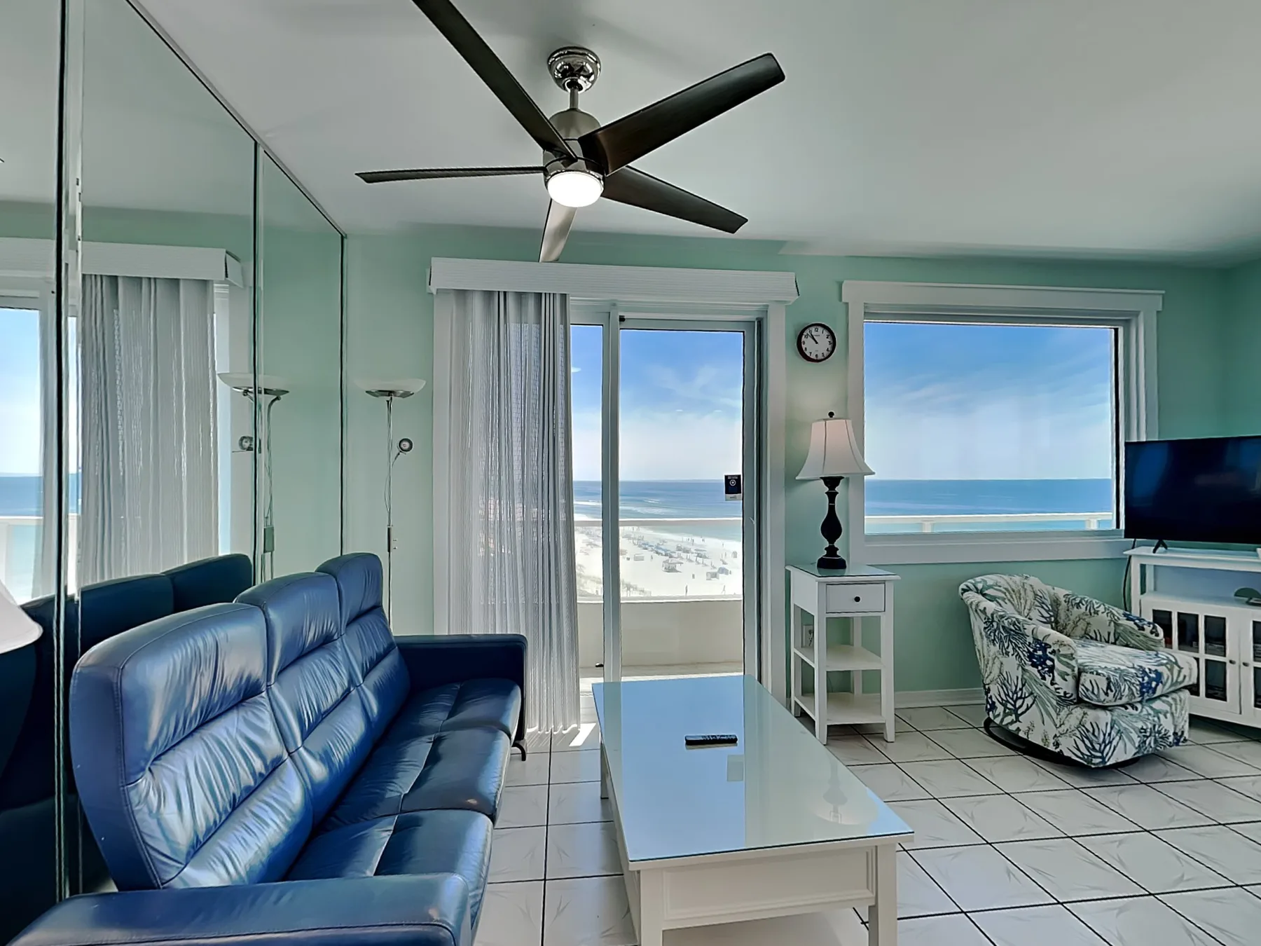 A bright living room with blue seating, a glass coffee table, and ocean views.