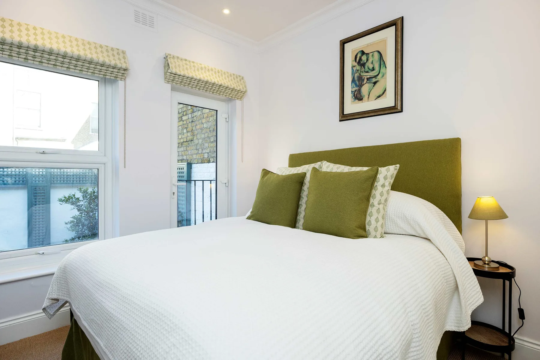 A bright bedroom with a neatly made bed, green headboard, and two tall windows.