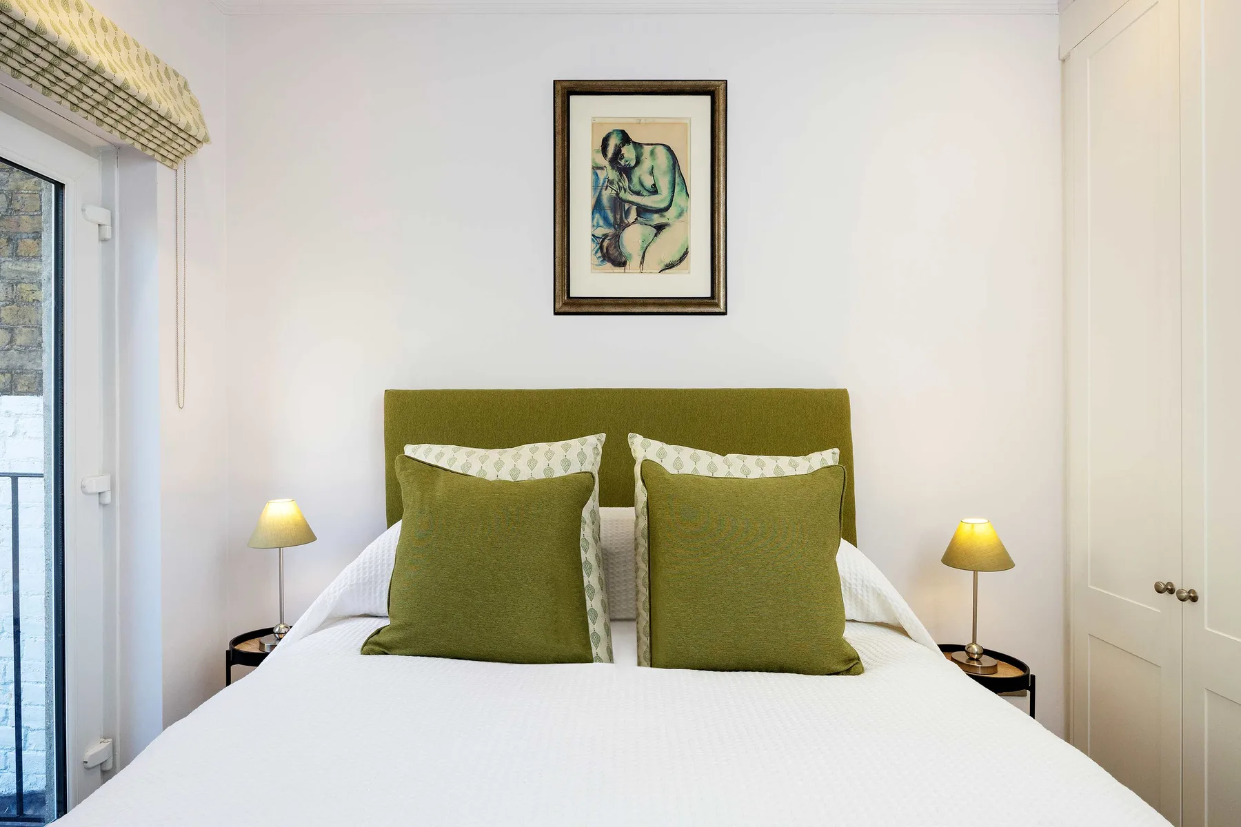 A bedroom with a bed, green headboard, and framed artwork above it.